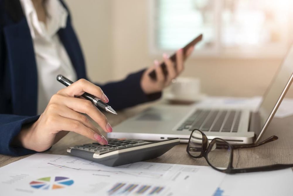 A person sits at a desk using a calculator and holding a smartphone, with financial documents, a laptop, and eyeglasses nearby, suggesting work related to finance or accounting.