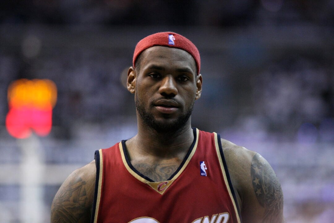 A basketball player wearing a red headband and a red Cleveland Cavaliers jersey stands on the court, looking forward with a neutral expression. The background is blurred with audience and stadium lights.