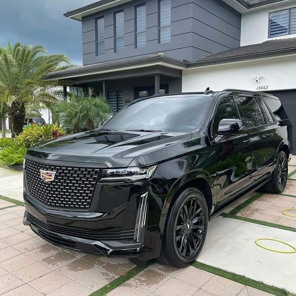 A shiny black Cadillac Escalade SUV is parked on a driveway in front of a modern two-story house with gray walls and palm trees in the yard.