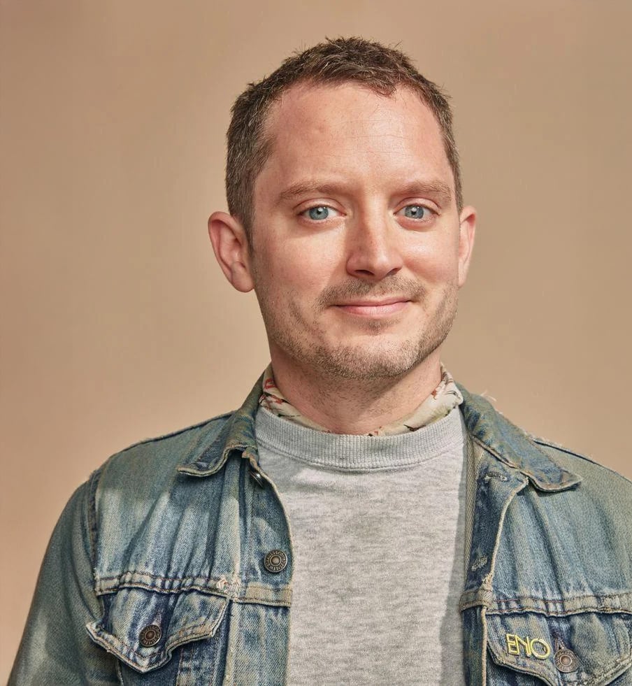 A smiling man with short brown hair wearing a denim jacket over a gray sweatshirt and a patterned scarf, posing against a plain beige background.