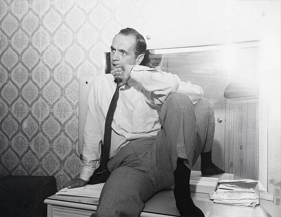 A man sits on a dresser in front of a mirror, resting his chin on his hand and gazing thoughtfully. He wears a white shirt, tie, and checkered pants. The room has patterned wallpaper and papers stacked nearby.