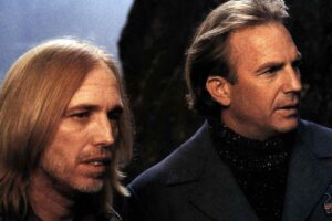 Two men with long hair stand side by side outdoors, looking into the distance. One has straight blond hair and a beard, while the other has short light brown hair and wears a dark coat with a US Mail patch.