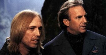 Two men with long hair stand side by side outdoors, looking into the distance. One has straight blond hair and a beard, while the other has short light brown hair and wears a dark coat with a US Mail patch.