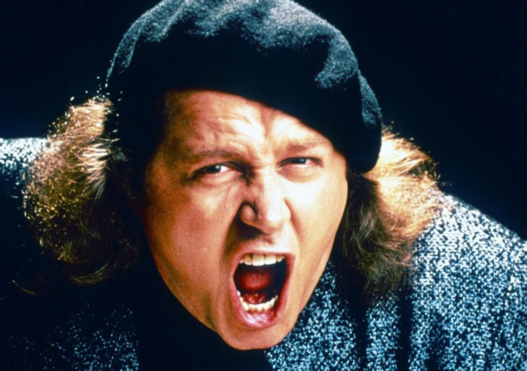 A person with long hair, wearing a black beret and a textured gray sweater, is shown mid-shout or yell with their mouth wide open against a dark background.