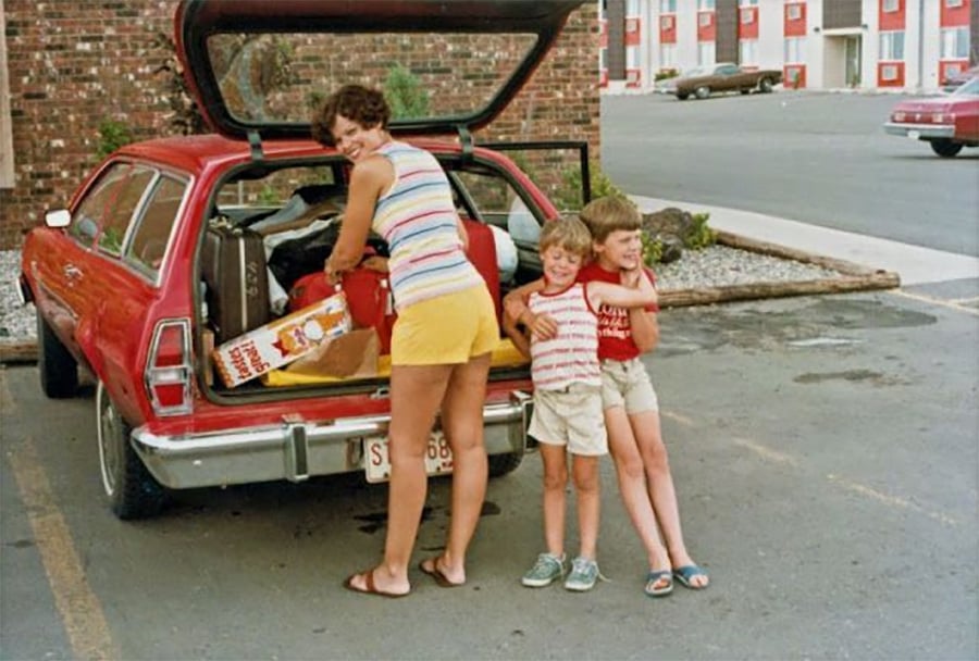 A woman smiles while packing a red station wagon with luggage. Two young boys stand nearby, hugging and smiling. The scene takes place in a parking lot with a brick building and parked cars in the background.