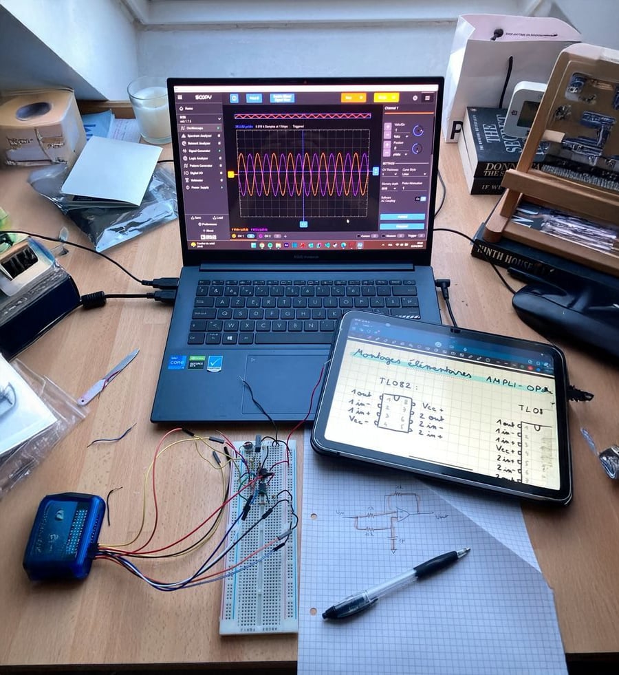 A cluttered desk with a laptop displaying waveform graphs, a tablet showing handwritten electronics notes, a breadboard with wires, electronic components, a blue device, a pen, and graph paper with a circuit diagram.