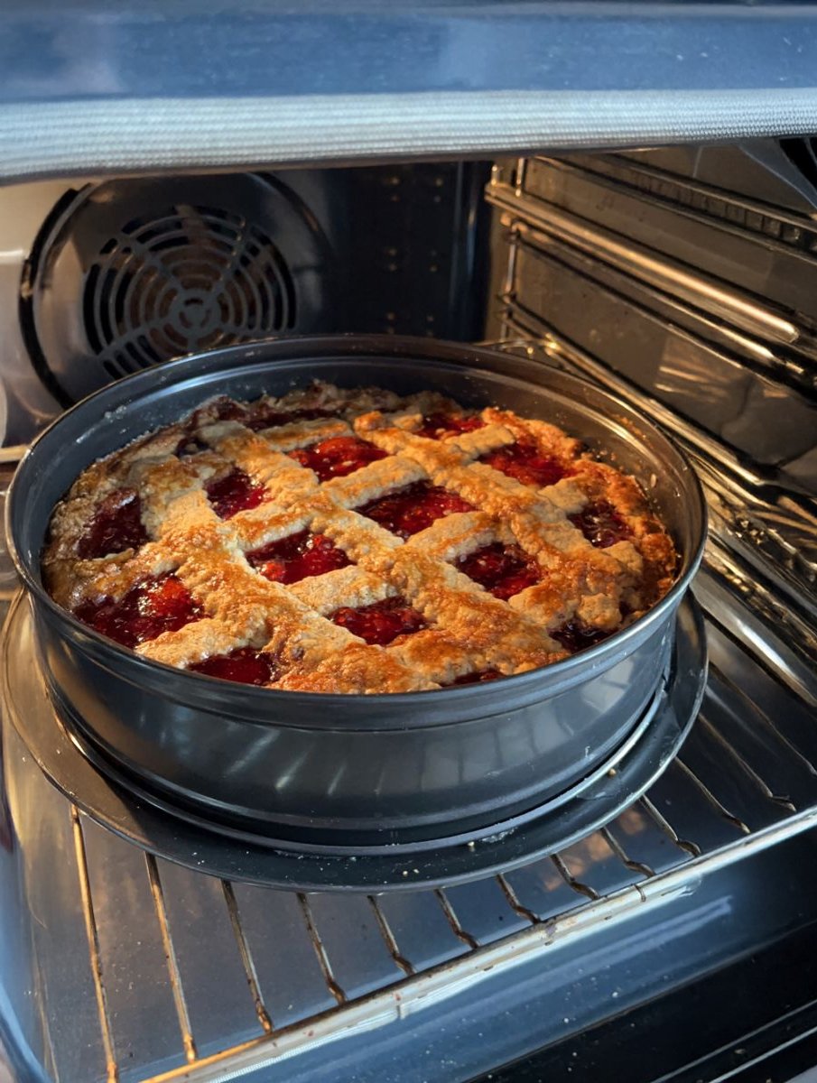 A homemade cherry pie with a golden-brown lattice crust is baking in an oven, resting on a round tray inside a circular pan.