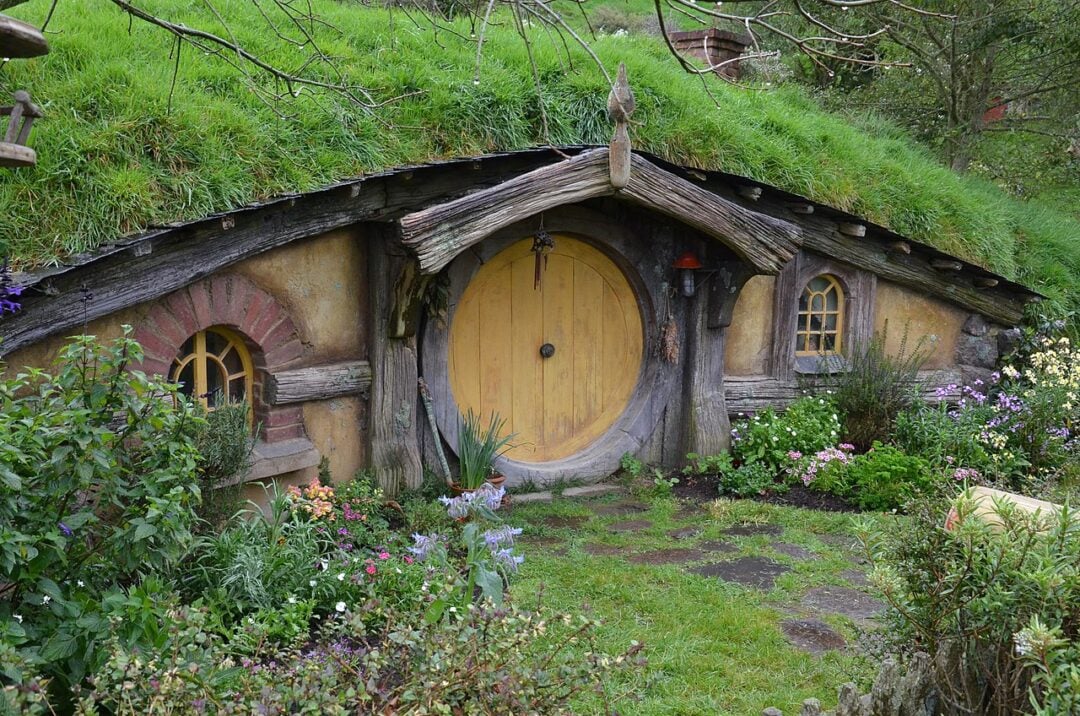 A charming hobbit house with a round yellow door, circular windows, and a roof covered in grass, surrounded by lush greenery and colorful flowers.