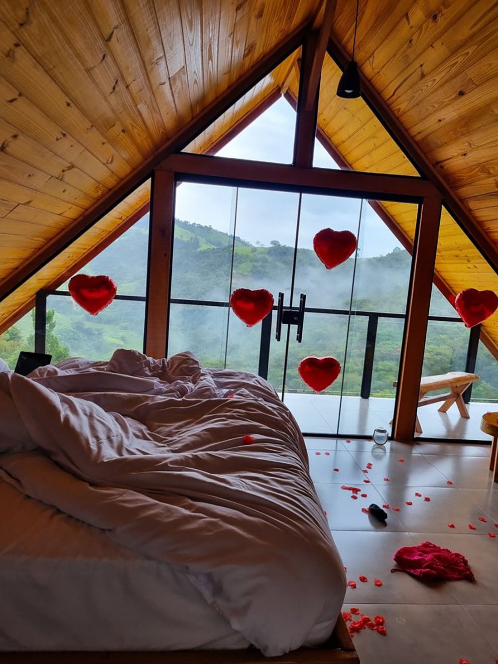 A cozy bed with white linens in a wooden cabin with a triangular ceiling. Red heart balloons and rose petals decorate the room. Large windows reveal a misty mountain view and lush green landscape outside.