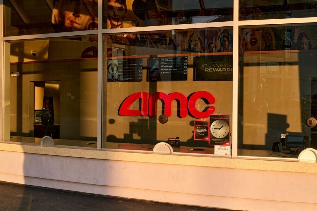 A glass ticket counter at an AMC movie theater displays the large red AMC logo. Movie posters and digital screens are visible inside, along with a clock and a sign about showtime restrictions on the window.