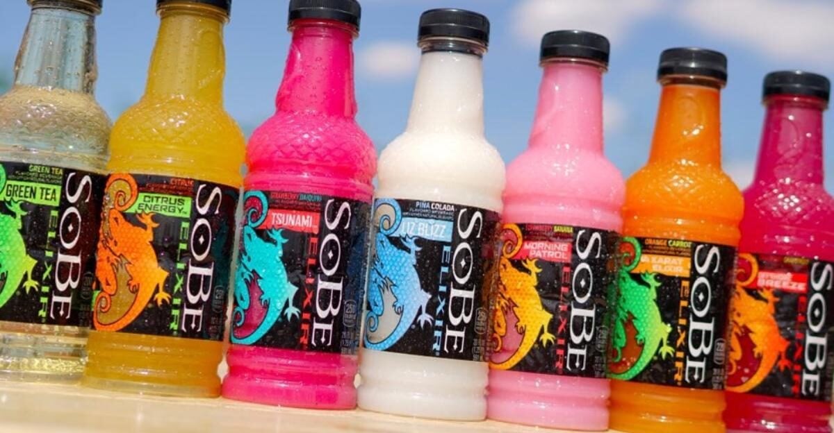Seven colorful SoBe drink bottles are lined up outdoors against a blue sky. Each bottle features a black label with a vibrant lizard graphic and different flavors and colors, including yellow, pink, blue, orange, and green.