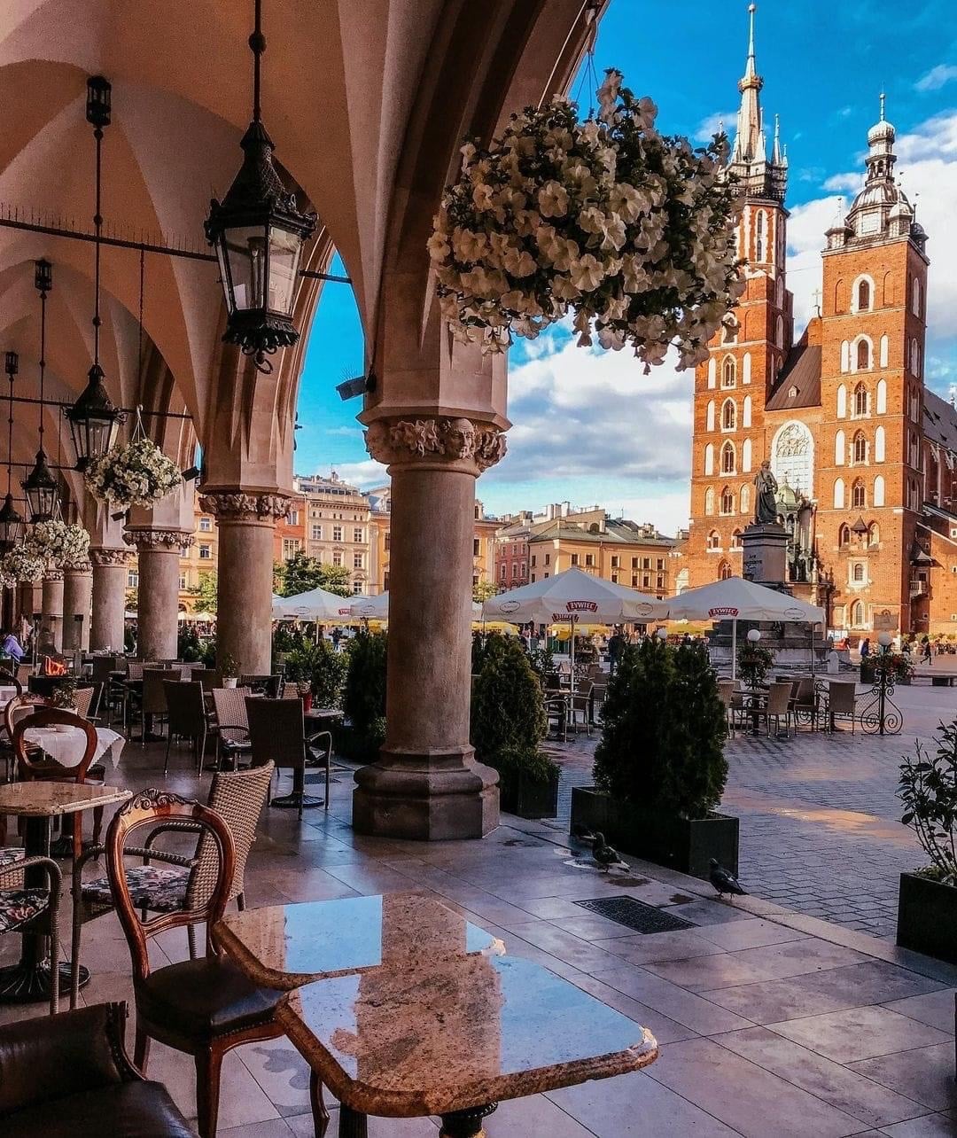 Outdoor café under arched columns with hanging flowers, overlooking a lively square and the St. Mary’s Basilica in Kraków, Poland, on a bright day with blue sky and scattered clouds.