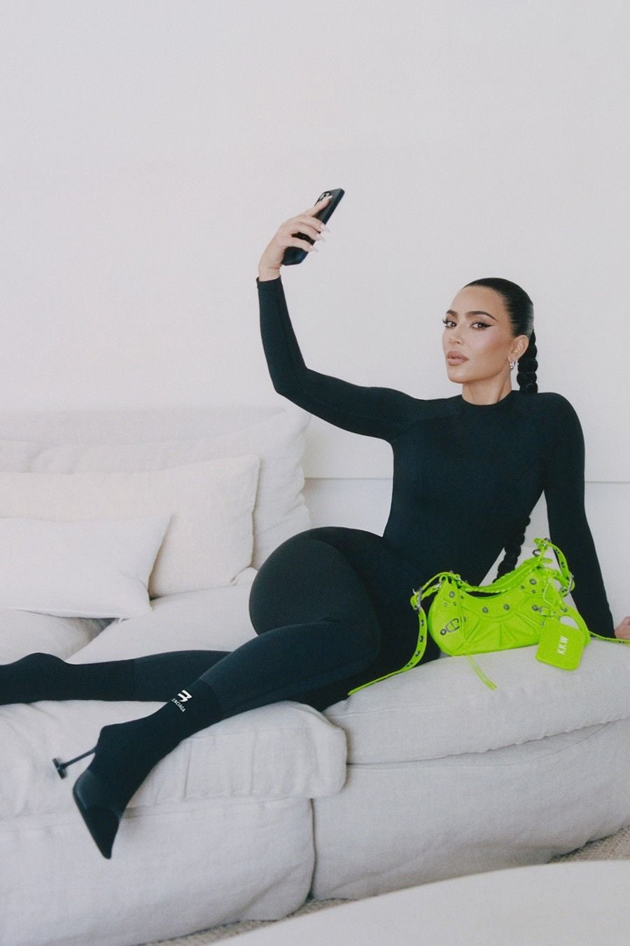 A woman in a fitted black outfit lounges on a white sofa, holding up a smartphone as if taking a selfie. A bright neon green handbag rests beside her. The background is minimal with white cushions.
