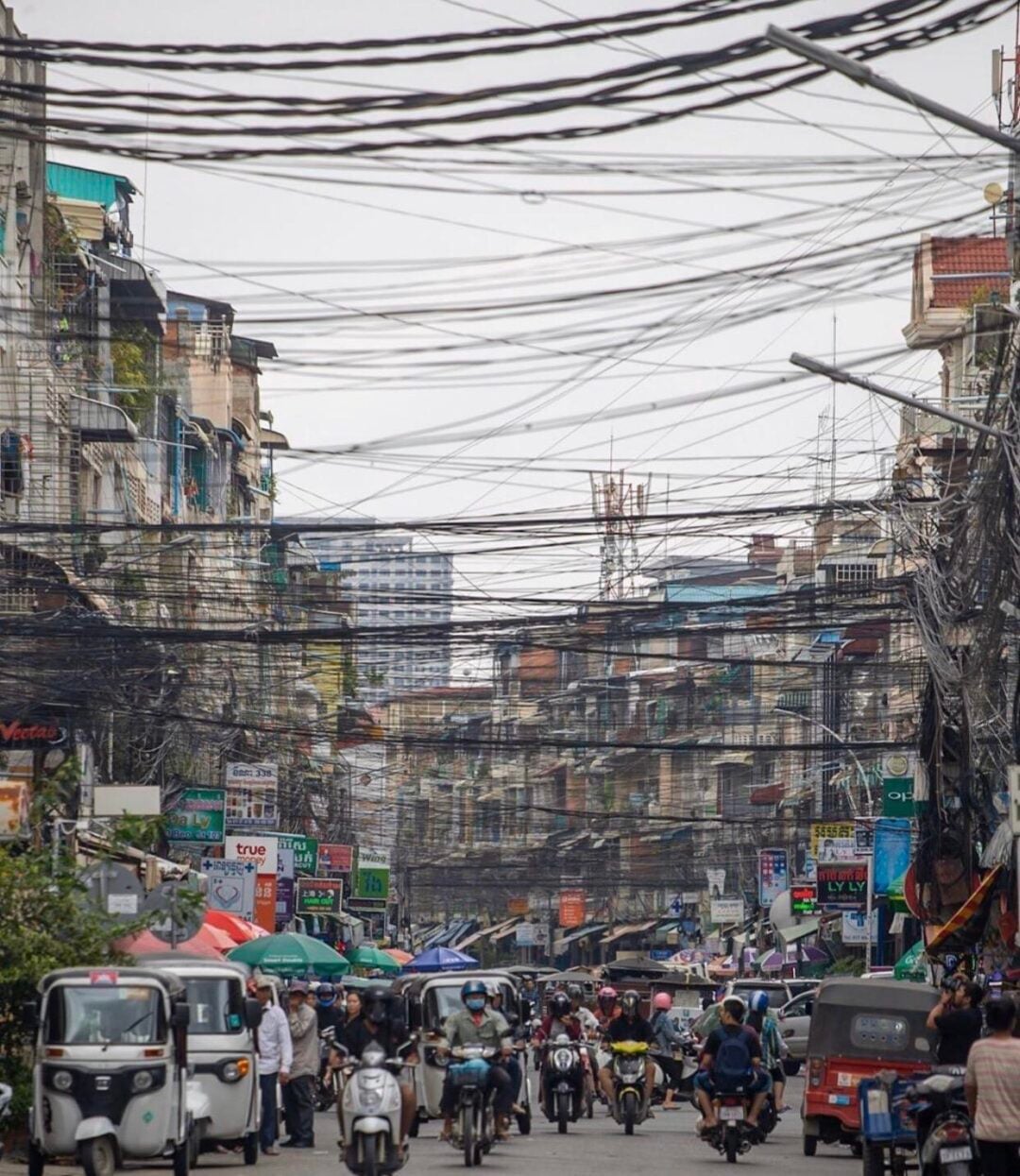 A busy urban street is filled with cars, tuk-tuks, and motorcycles. Numerous overhead electrical wires crisscross above, creating a tangled web between densely packed buildings. People and shops line both sides of the street.