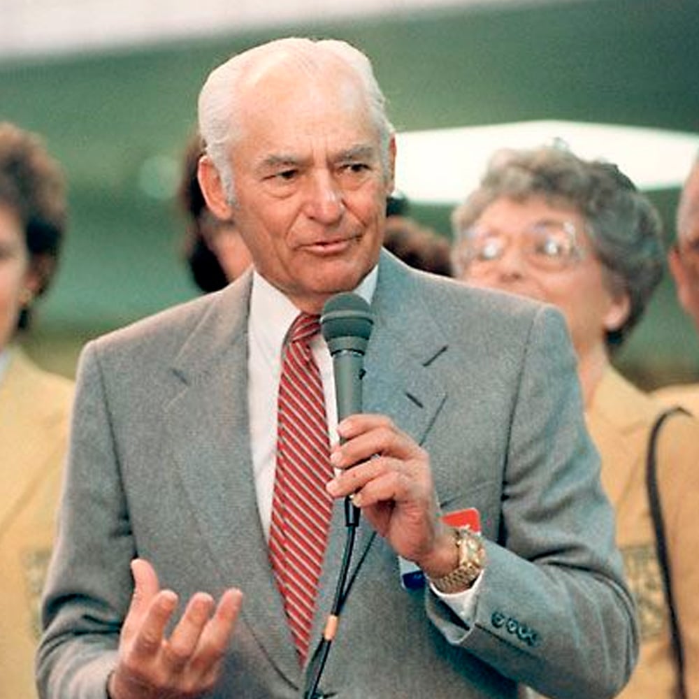 An older man in a gray suit and red tie speaks into a microphone, gesturing with his hand. He is standing in front of a group of people, some wearing yellow jackets, in an indoor setting.