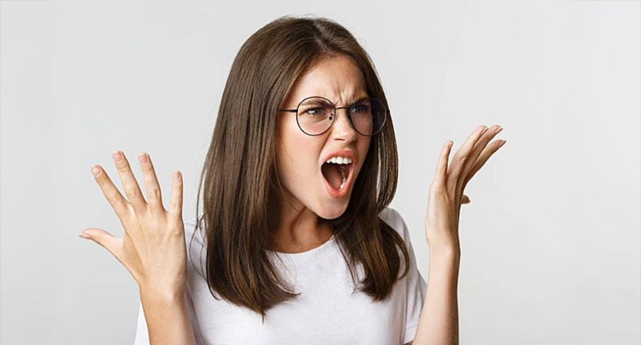 A young woman with long brown hair and glasses is wearing a white t-shirt, raising her hands with fingers spread and mouth open, appearing frustrated or upset against a plain light background.