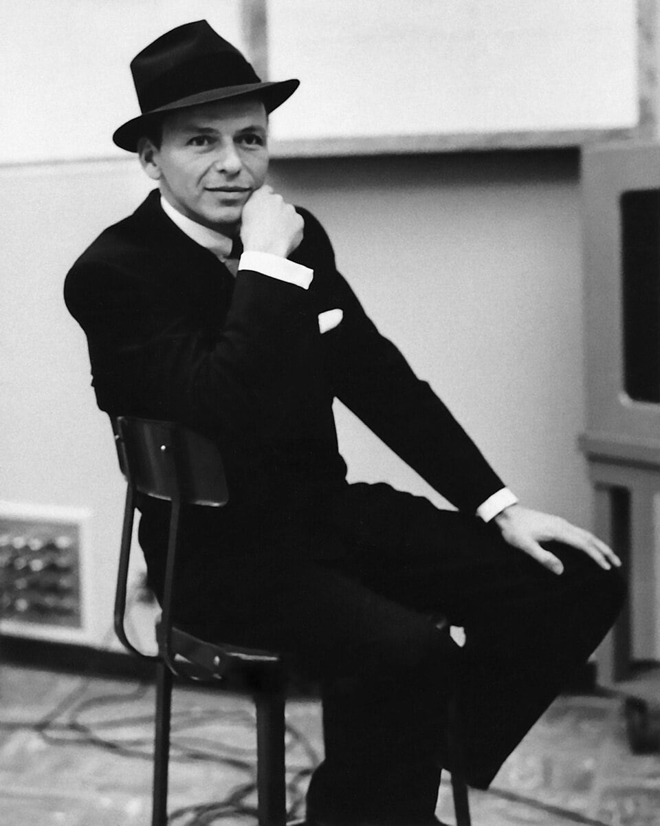 A man in a dark suit, tie, and fedora hat sits sideways on a chair, resting one arm on the backrest and the other on his knee, looking at the camera with a relaxed expression in a vintage indoor setting.
