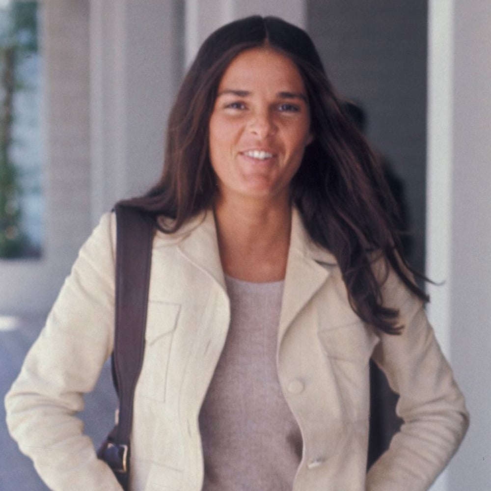 A woman with long dark hair, wearing a beige jacket over a light top, stands outside near a building and smiles at the camera, carrying a dark shoulder bag.