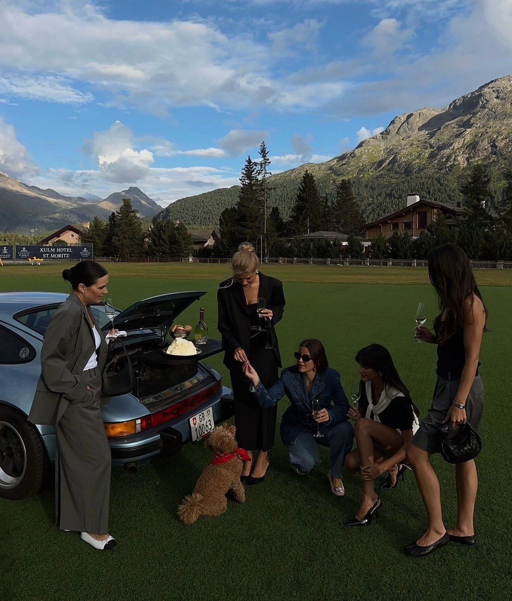 Five stylish women stand and sit near a blue vintage Porsche parked on grass, sharing drinks and snacks by the open trunk, with a small dog nearby. Green mountains and blue skies fill the scenic background.