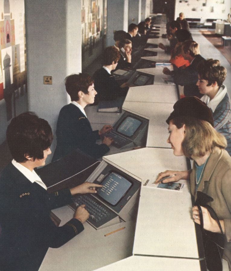 People sit on both sides of a long counter using vintage computer terminals, likely in an office or travel booking setting. Women in uniforms assist customers seated across from them. The scene appears to be from the mid-20th century.