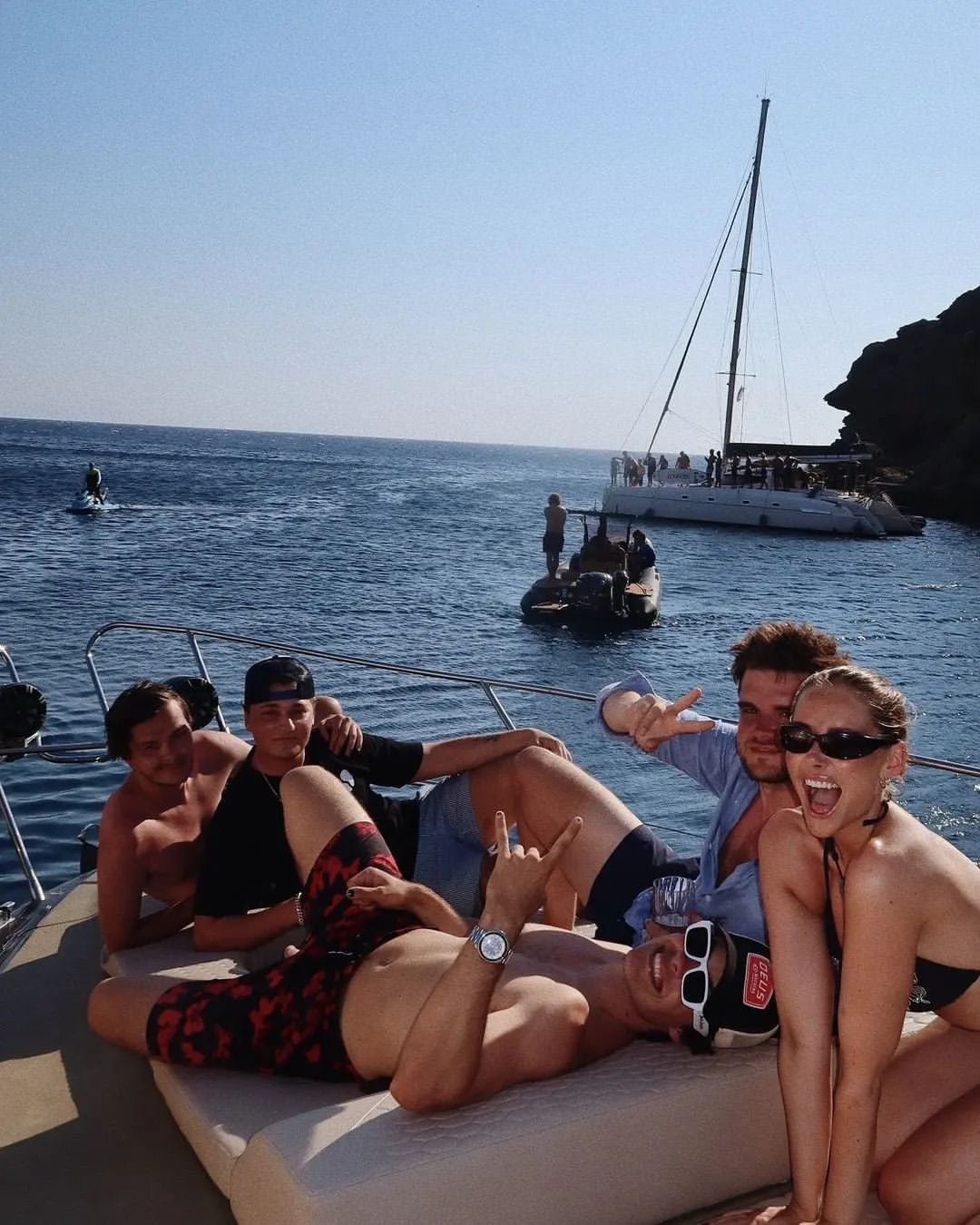 Four young adults relax and pose playfully on a yacht, smiling and making hand signs. Behind them, a sailboat and rocky coastline are visible under a clear, sunny sky.