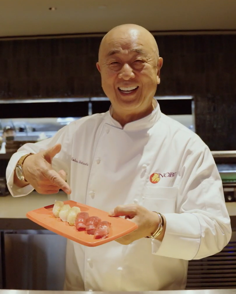 A smiling chef in a white coat stands indoors, holding a rectangular orange platter with pieces of sushi and pointing at them. The chef’s coat has a Nobu logo on it.
