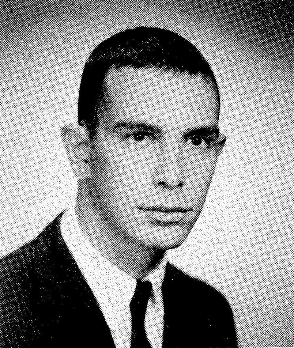 Black and white portrait of a young man with short hair, wearing a suit, white shirt, and tie, facing forward with a neutral expression.