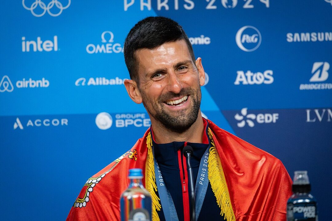 A smiling athlete draped in a red and gold flag sits at a press conference table with a Paris 2024 backdrop featuring various sponsor logos. A medal hangs around his neck and water bottles are on the table.