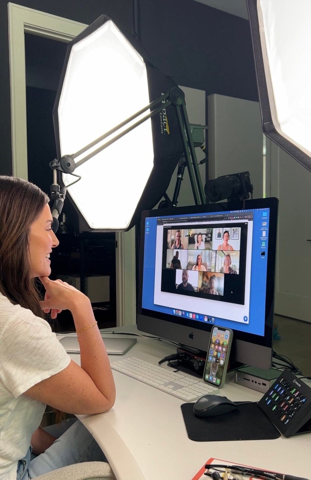 A woman sits at a desk in front of a computer monitor, participating in a video call with several people. Two large studio lights are set up behind the screen, and a smartphone is propped up beside the keyboard.
