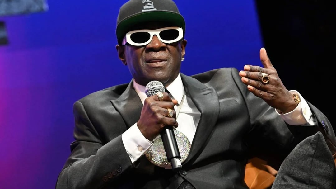 A man in a dark suit, white sunglasses, and a black hat gestures while speaking onstage, wearing a large clock necklace and multiple rings, against a purple and blue background.