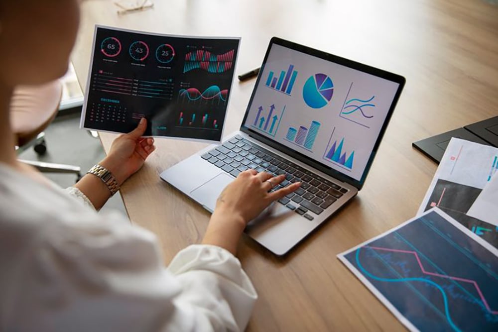A person sits at a desk, viewing colorful charts and graphs on a laptop screen and holding a printed data report with similar visualizations. Other data sheets are spread on the wooden desk.