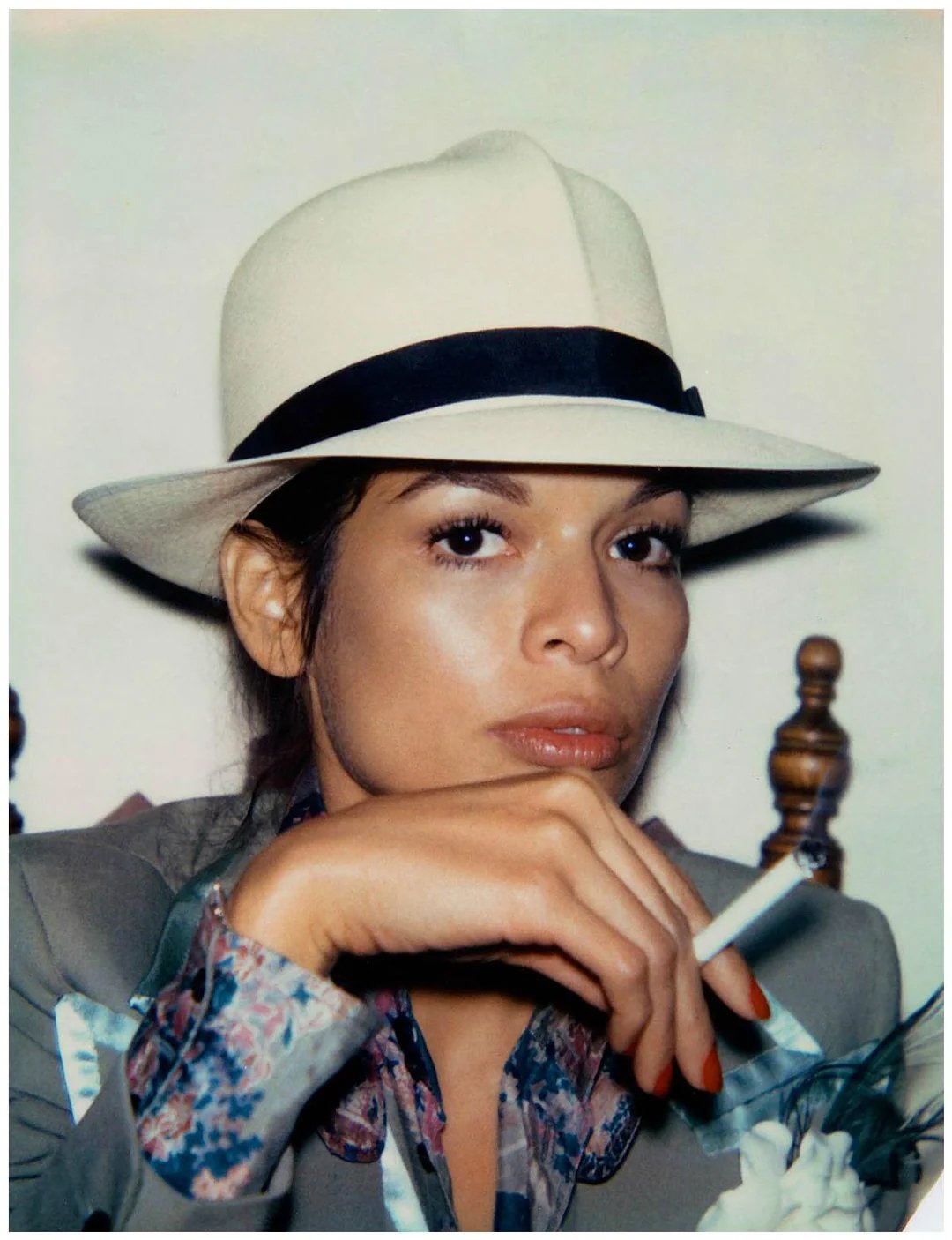 A person wearing a cream-colored fedora and floral shirt holds a cigarette, gazing confidently at the camera with a neutral expression.
