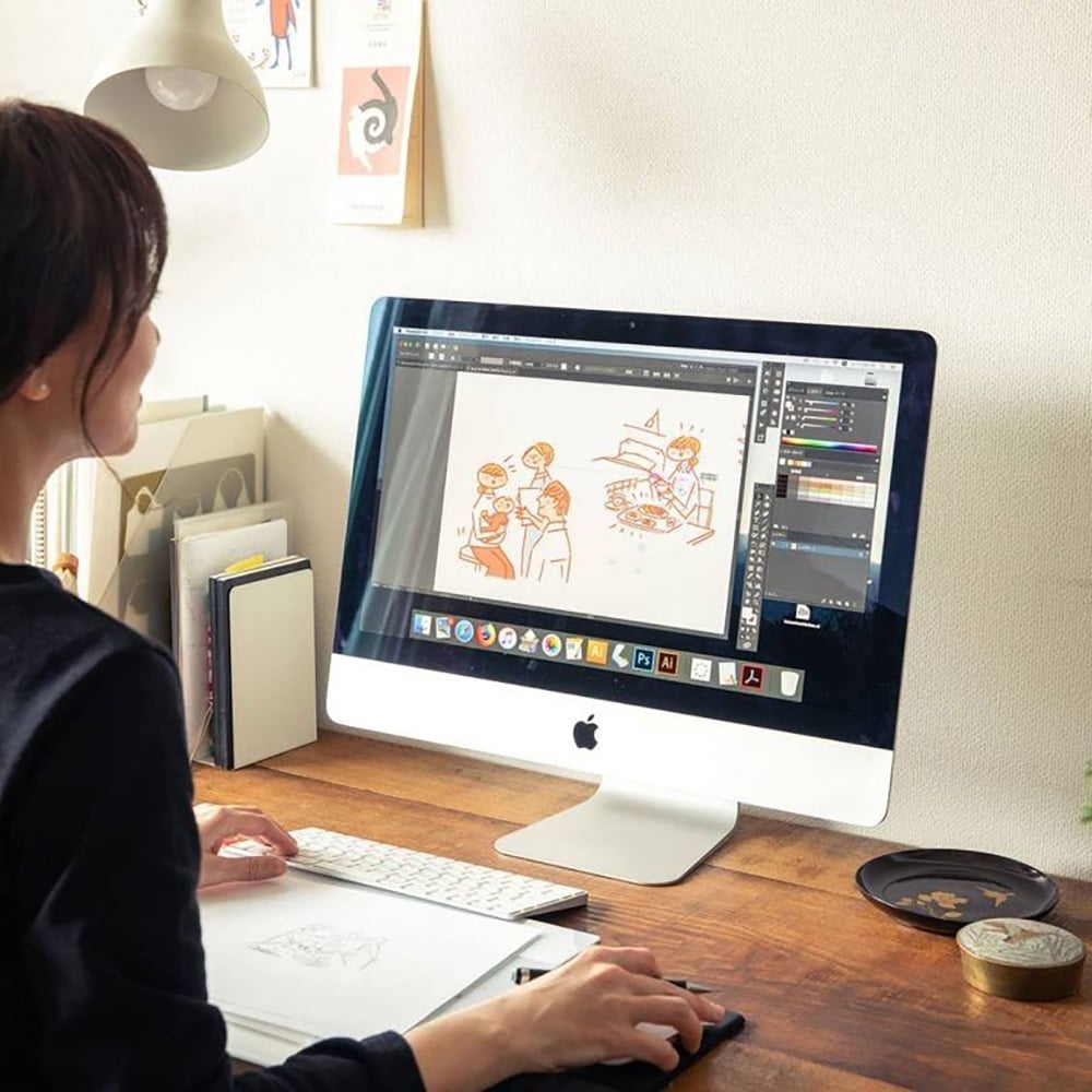 A person sits at a desk working on a drawing of people and food on a large iMac screen using illustration software, with art supplies and sketchbooks nearby in a well-lit workspace.