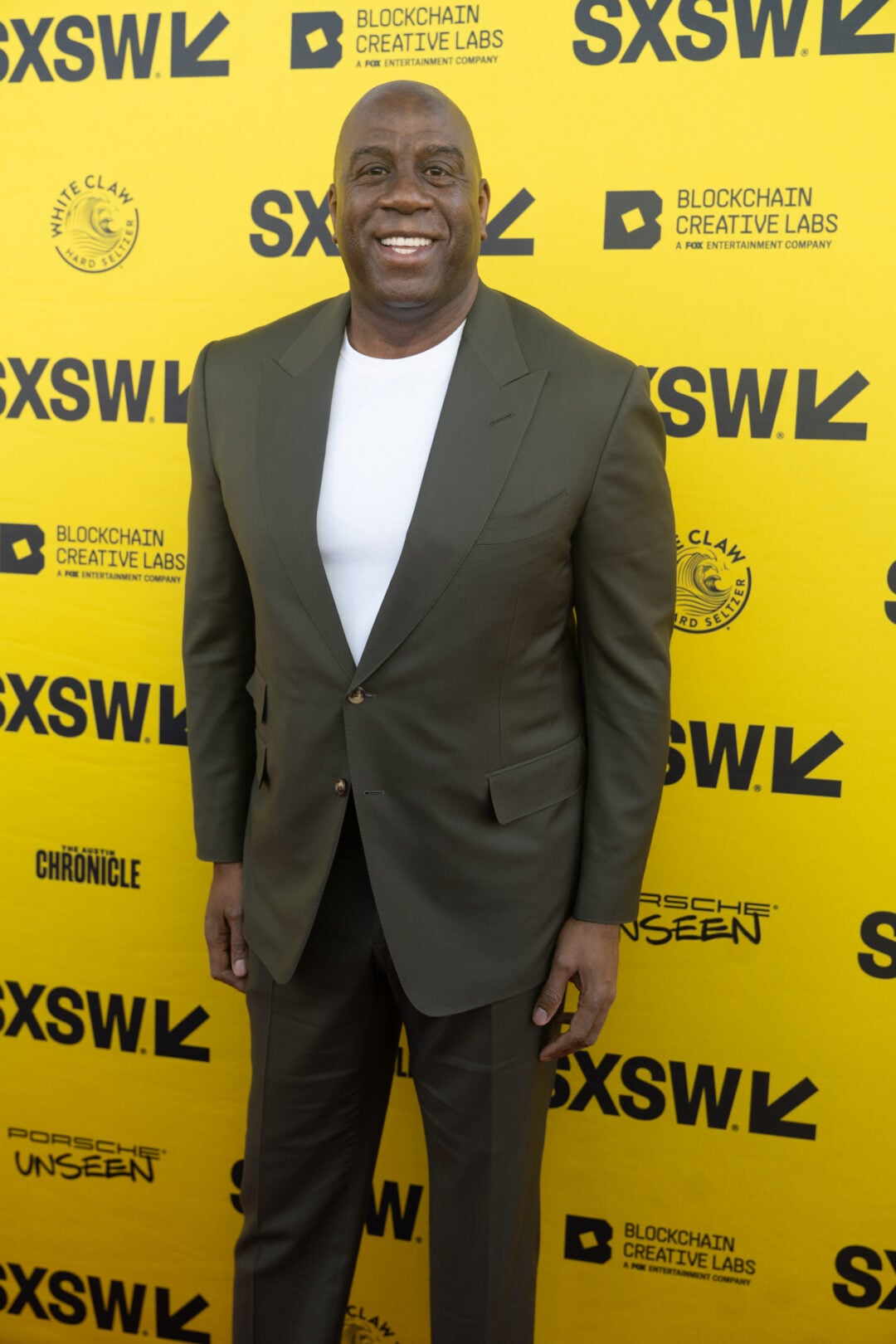 A smiling man in a green suit and white shirt stands in front of a bright yellow SXSW backdrop featuring various sponsor logos.