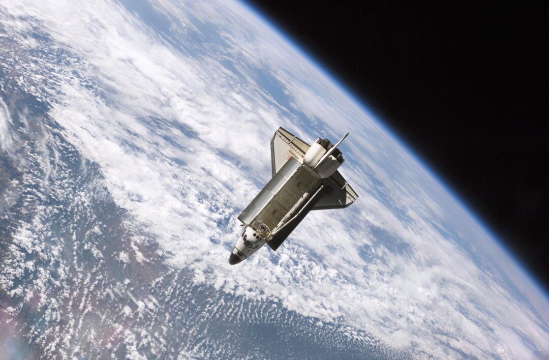 A space shuttle orbits above Earth, with the planet’s blue surface and white clouds visible below. The shuttle’s cargo bay doors are open, and the blackness of space curves along the image’s top edge.