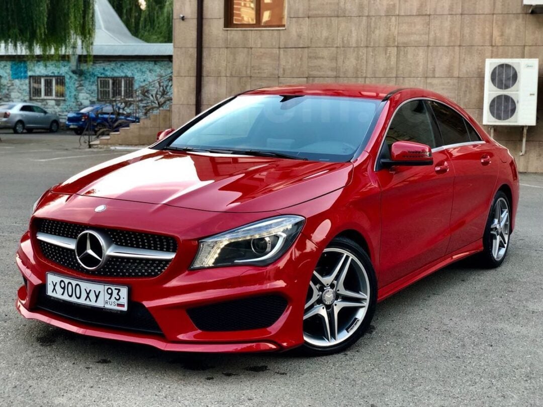 A shiny red Mercedes-Benz sedan is parked on a street in front of a beige building, with reflective windows and a wall-mounted air conditioner unit in the background. The car has Russian license plates.