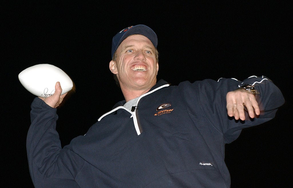 A smiling man in a navy blue sports jacket and cap throws a white football at night, with his arm cocked back and his mouth open in excitement.
