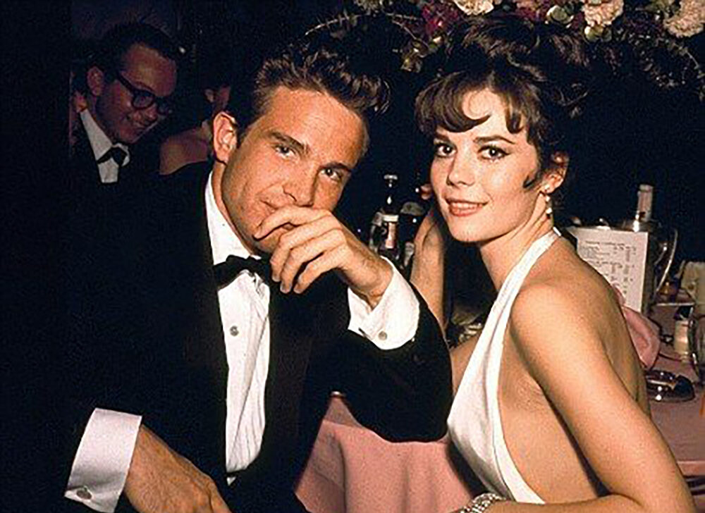 A man in a suit and a woman in a white halter dress sit closely together at a formal event, smiling at the camera. The background shows other guests and floral decorations.