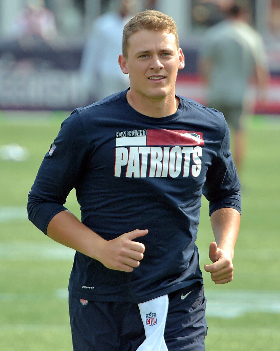 A football player in a navy blue long-sleeve shirt with "PATRIOTS" on the front is jogging on a field. He appears to be warming up, with other people and blurred background elements visible behind him.