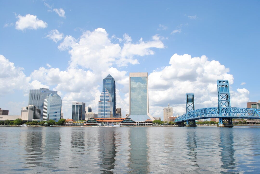 A city skyline with modern skyscrapers and a bright blue bridge over a calm river under a partly cloudy sky. The buildings and bridge are reflected in the water.