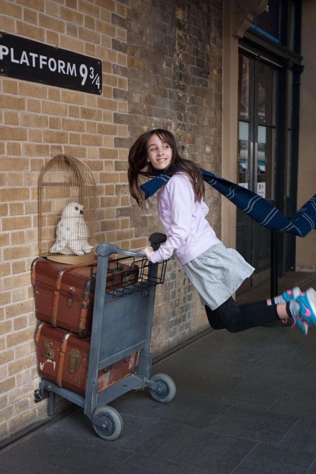 A girl wearing a scarf and sneakers poses as if she is running through the wall at Platform 9 3/4, pushing a trolley with suitcases and a caged owl, inspired by Harry Potter, at King's Cross Station.