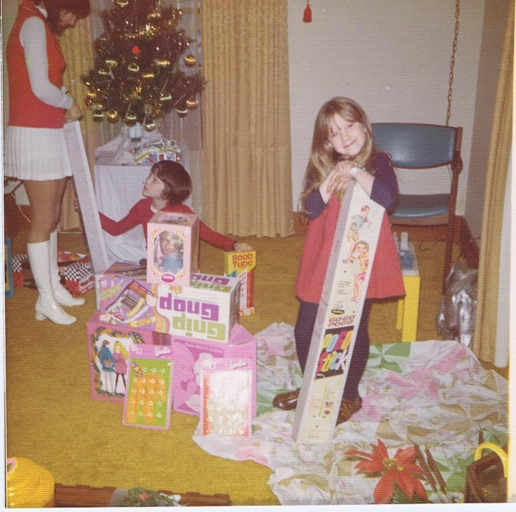 Two children open presents near a Christmas tree, surrounded by toys and wrapping paper. One child smiles, holding a long box; a woman in a white skirt and boots stands beside them. Festive decor is visible in the background.