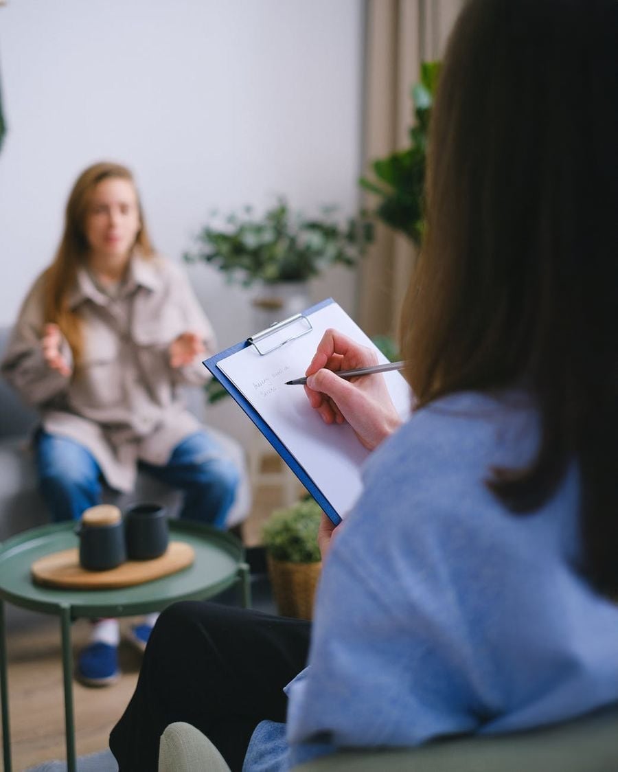 A person sits with a clipboard and pen, taking notes, while another person talks with expressive hand gestures in a cozy indoor setting, suggesting a therapy or counseling session.