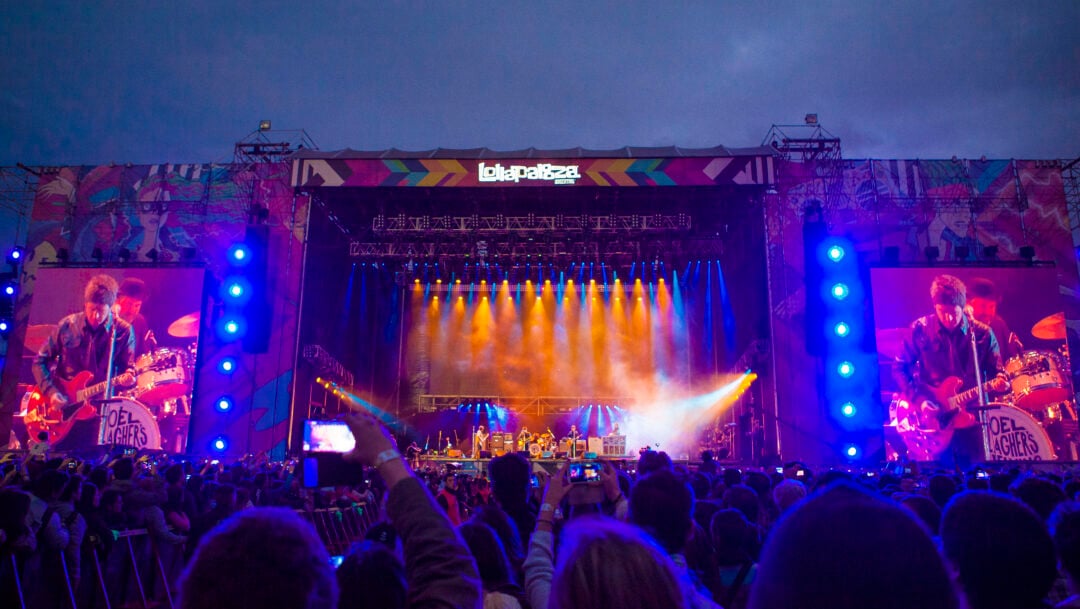 A large crowd watches a live band perform on a brightly lit outdoor stage at night during Lollapalooza, with colorful lights and big screens displaying musicians on either side of the stage.