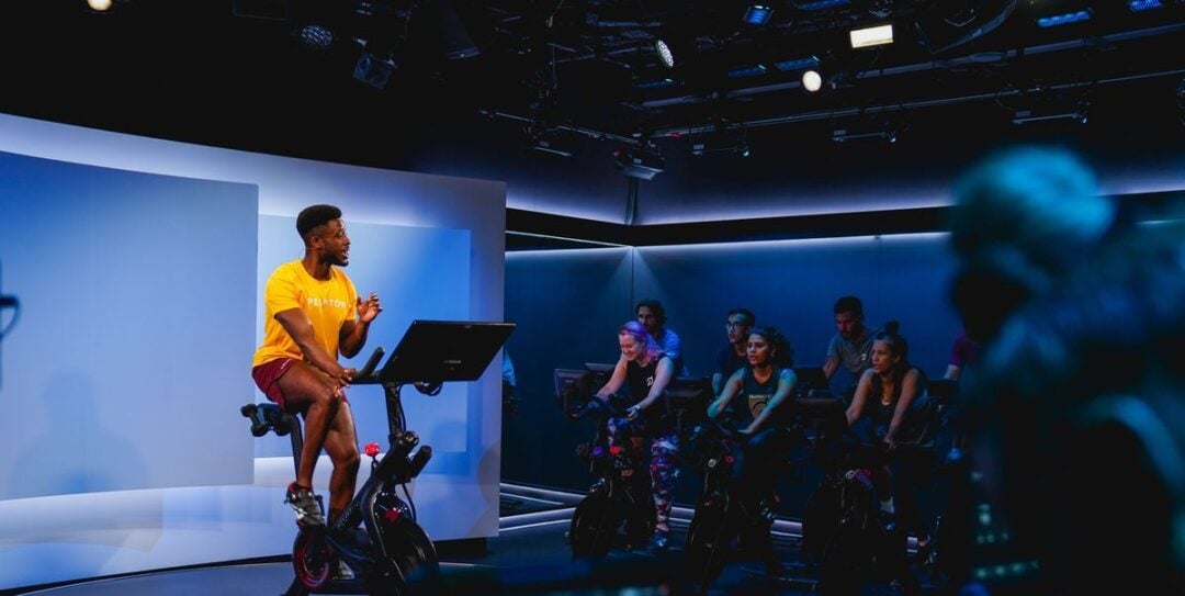 A fitness instructor on a stationary bike leads a spin class in a dimly lit studio, speaking to a group of people exercising on bikes. The room has blue lighting and a modern atmosphere.