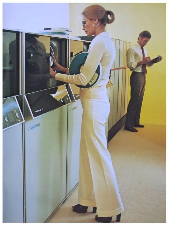 A woman in 1970s-style clothing loads a large data reel onto a computer, while a man in the background reads paperwork near a row of vintage mainframe machines in an office setting.