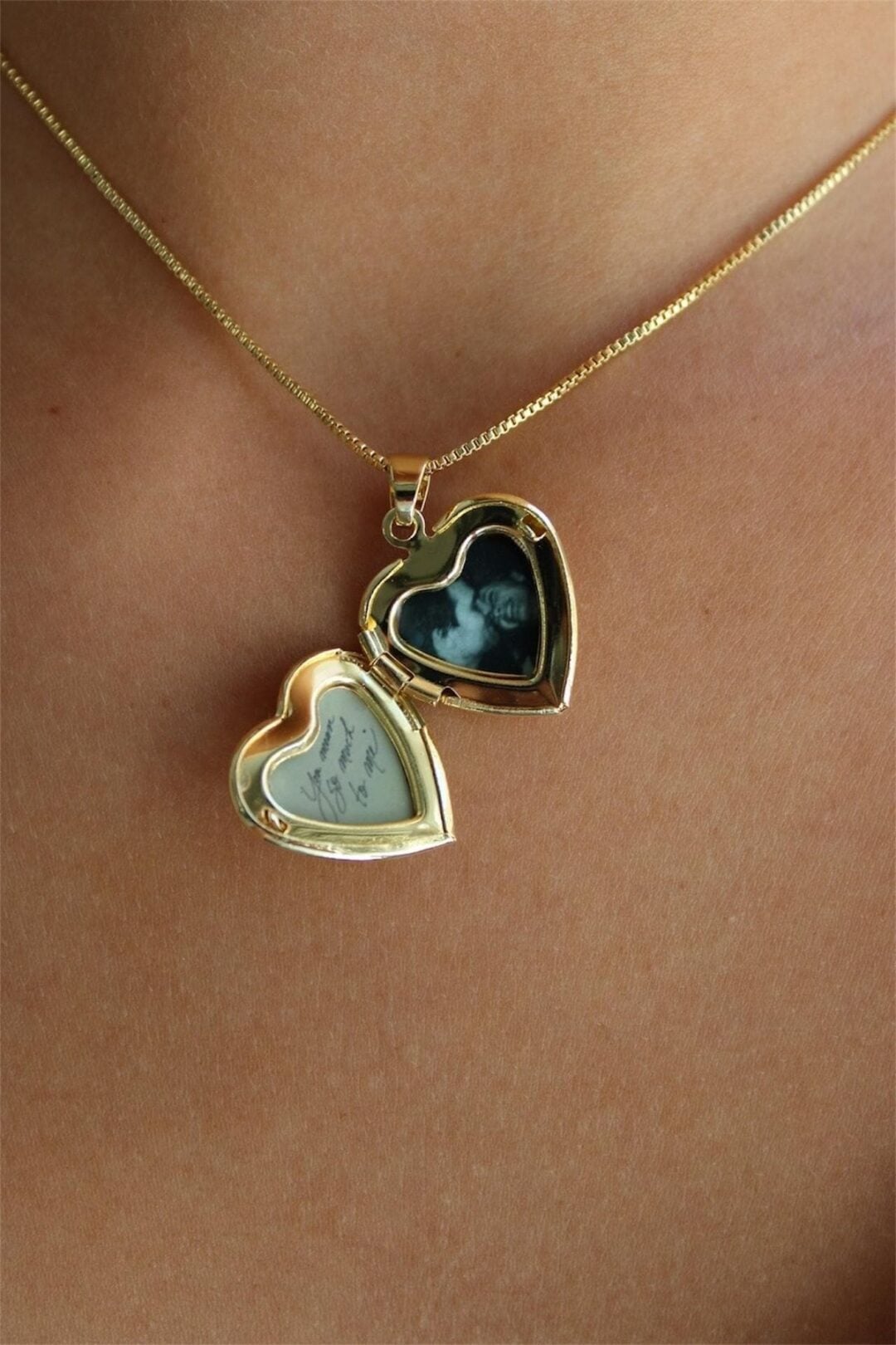 A close-up of a gold heart-shaped locket necklace, open to reveal a small black-and-white photo on one side and a handwritten note on the other, worn against bare skin.
