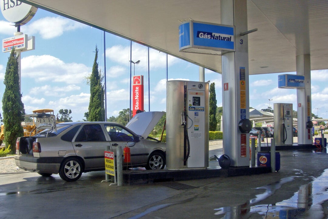 A silver car with its hood open is parked at a gas station pump labeled "Gás Natural." The station has multiple fuel pumps, signs, and a clear sky with clouds in the background.