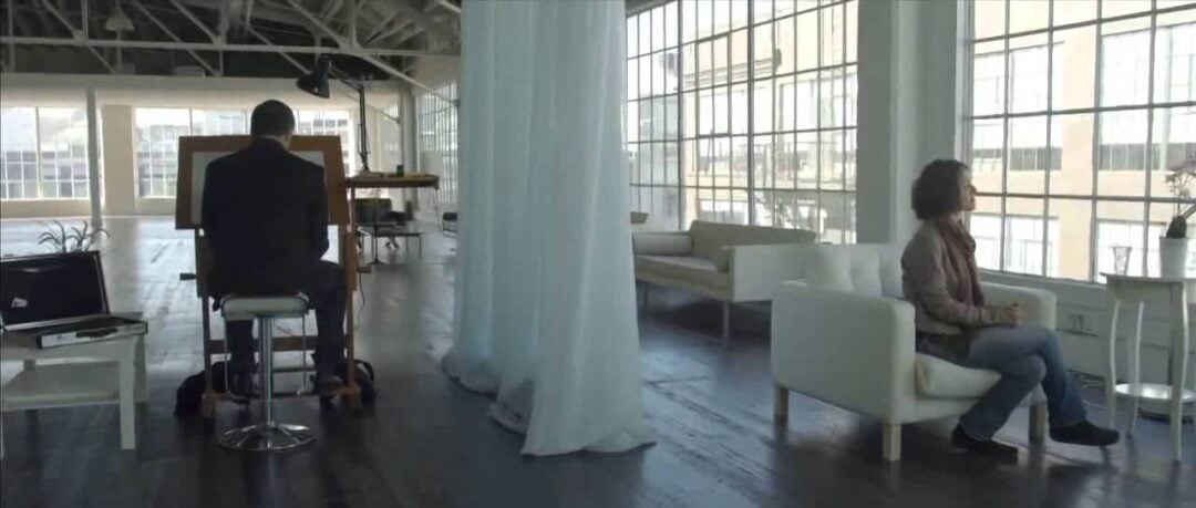 A man sits at an easel drawing while a woman sits on a white couch across a spacious, sunlit room with large windows and sheer curtains. The setting appears to be an art studio or loft.