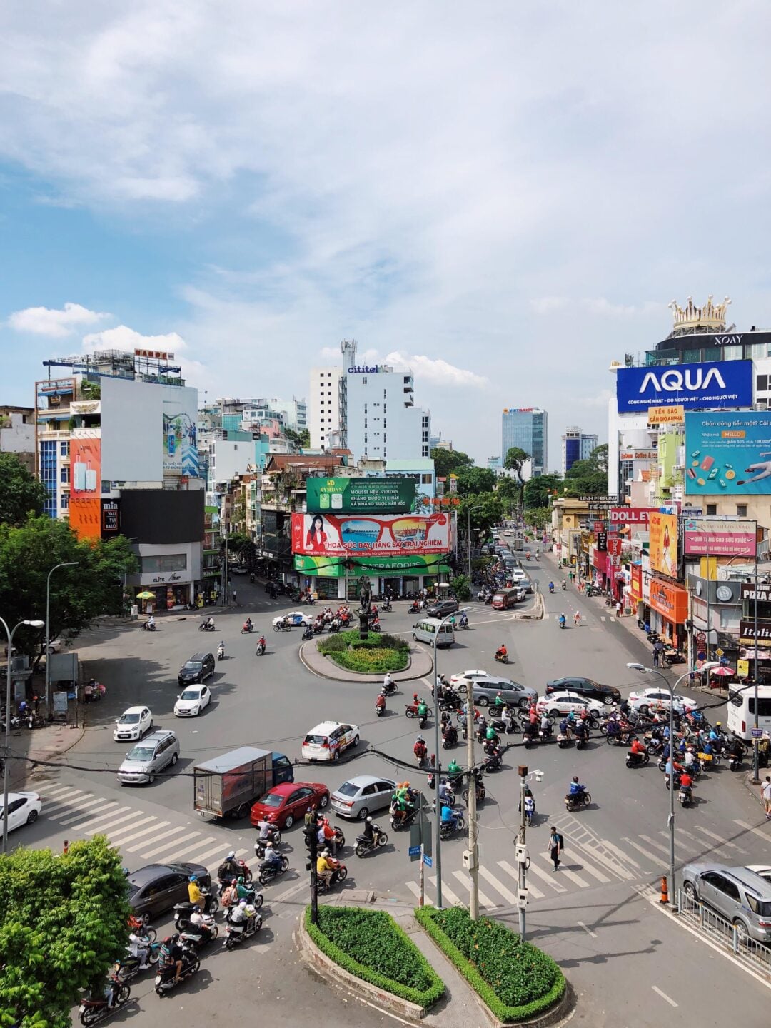 A busy urban roundabout with cars, buses, and many motorbikes navigating the streets, surrounded by tall buildings, billboards, and greenery under a partly cloudy sky.